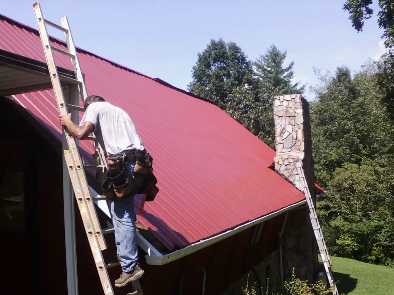 Roofer on a ladder beside a red metal roof and stone chimney — Soffit & Fascia Repair by Roofing Brothers in Wrightwood